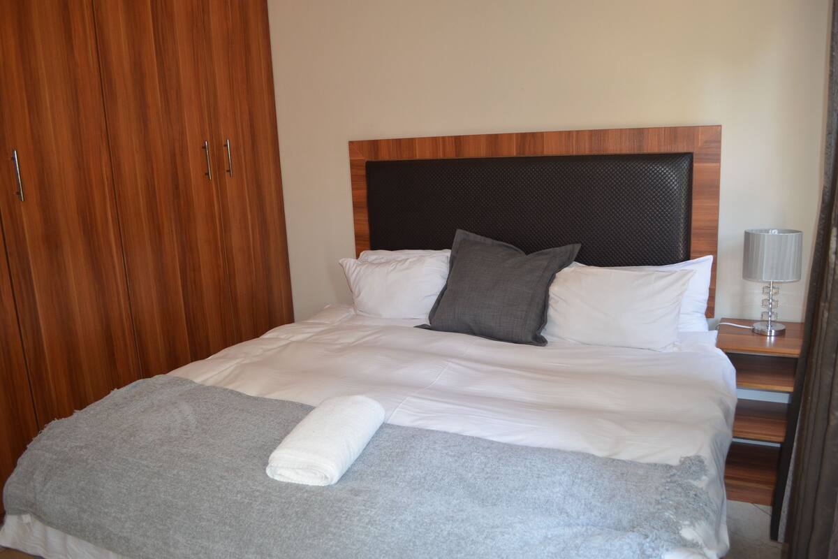 Cama bem arrumada com roupa de cama branca e cinza, cabeceira de madeira e uma pequena mesa de cabeceira com abajur.