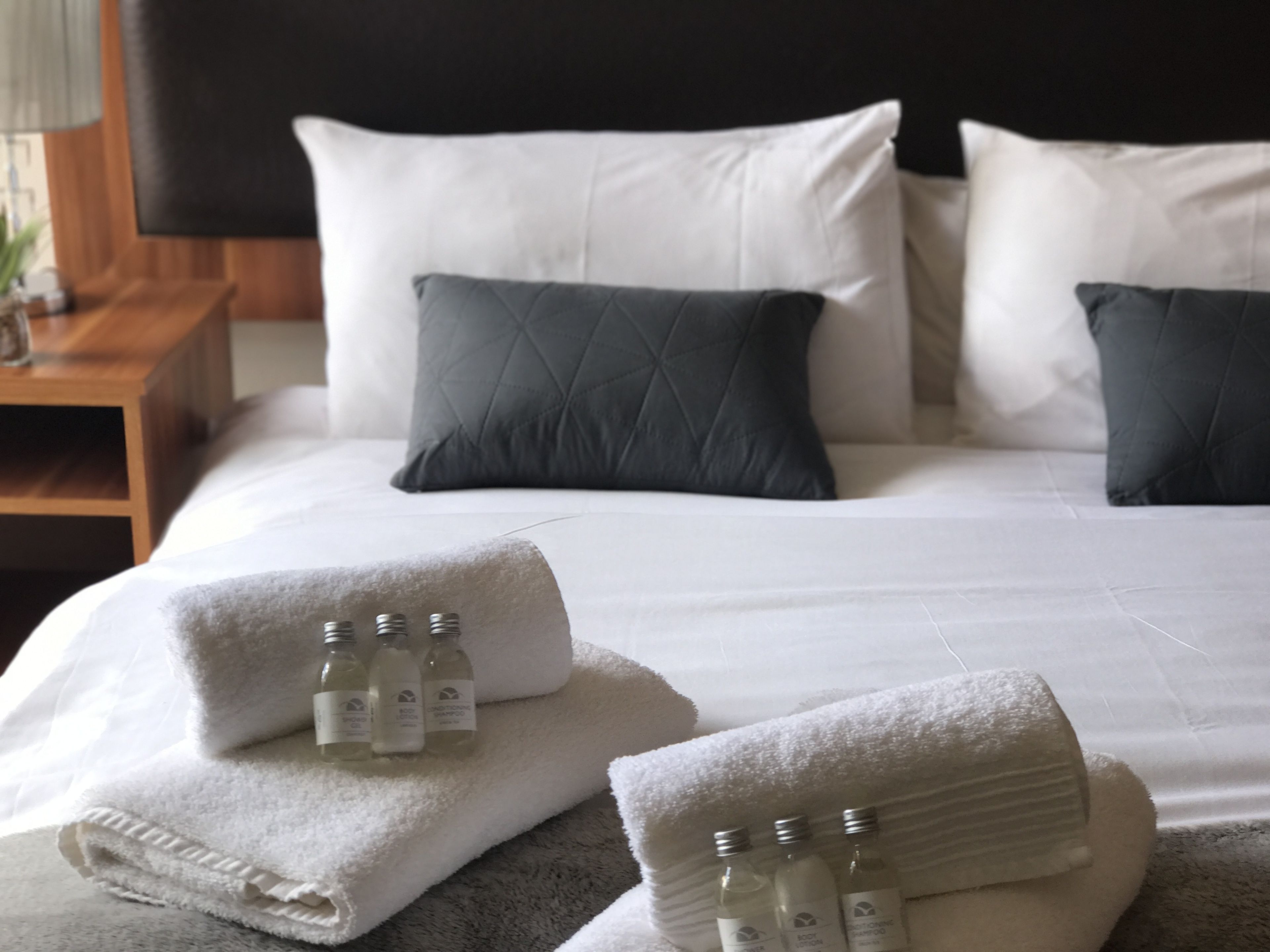A neatly made bed with white and dark grey pillows, a grey headboard, and towels with bottles of toiletries.