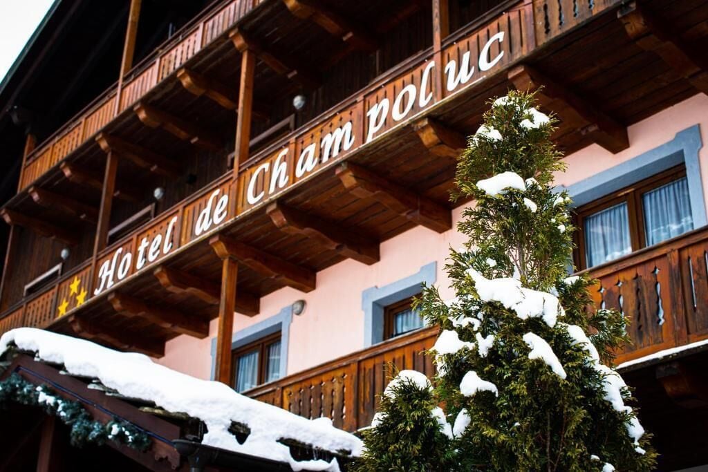 A hotel with a wooden facade and balconies, snow on the roof and trees, and a sign reading 'Hotel de Cham de Ross.'