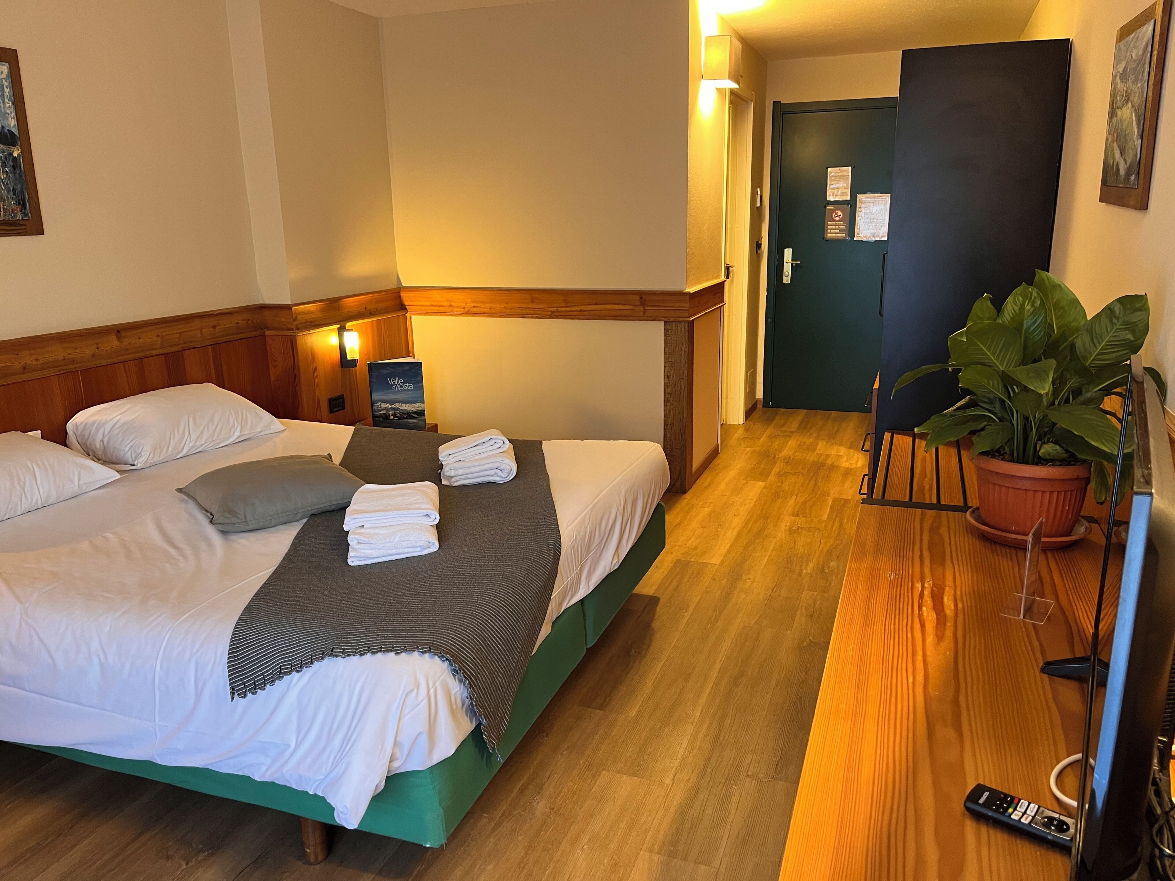 Ein Hotelzimmer mit einem Bett, Kissen, einem gefalteten Handtuch, einem Nachttisch mit Lampe, einer Topfpflanze und einer Tür mit einem Schild.