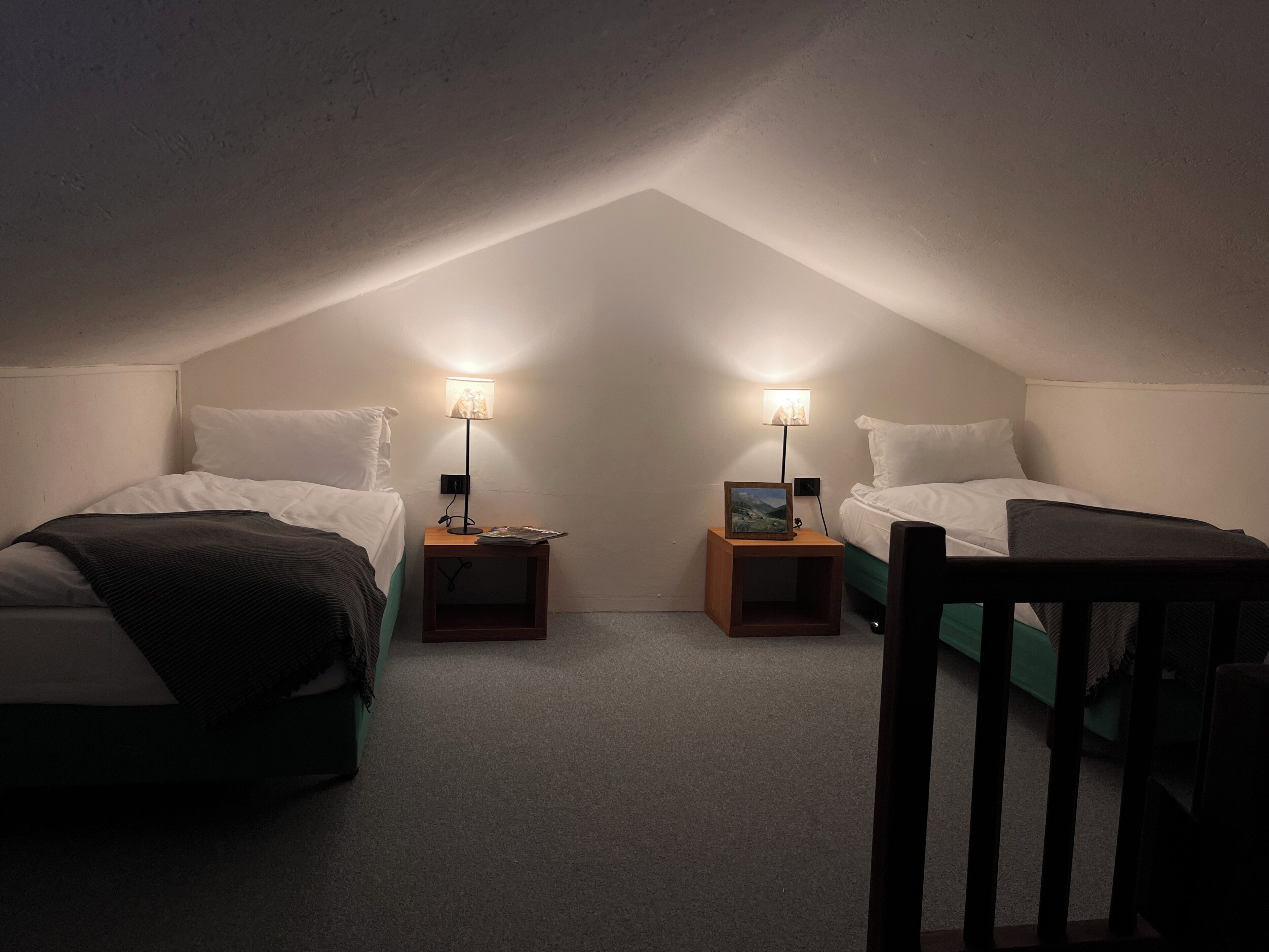 Ein kleines Schlafzimmer im Dachgeschoss, schwach beleuchtet, mit zwei Betten, jeweils mit Nachttisch und Lampe.