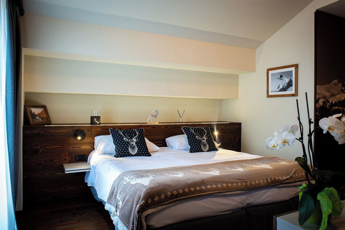 A neatly made bed with a patterned bedspread, two pillows with deer designs, and a bedside table with a framed picture and a small vase.