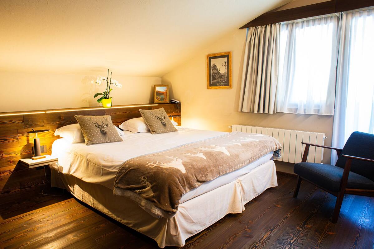 A hotel room with a large bed, a nightstand, a chair, a window with curtains, a radiator, and a framed picture on the wall.