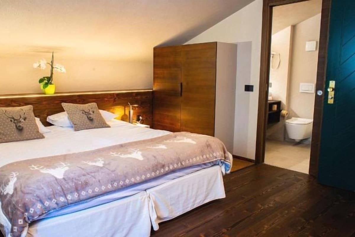 A bedroom with a bed, nightstands, a wooden wardrobe, and a bathroom visible through an open door.