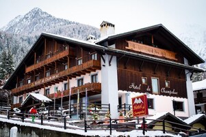 Un hotel de estilo chalet de madera con balcones, un letrero que indica que es el Hotel de Champoluc, y una vista de montañas nevadas al fondo.