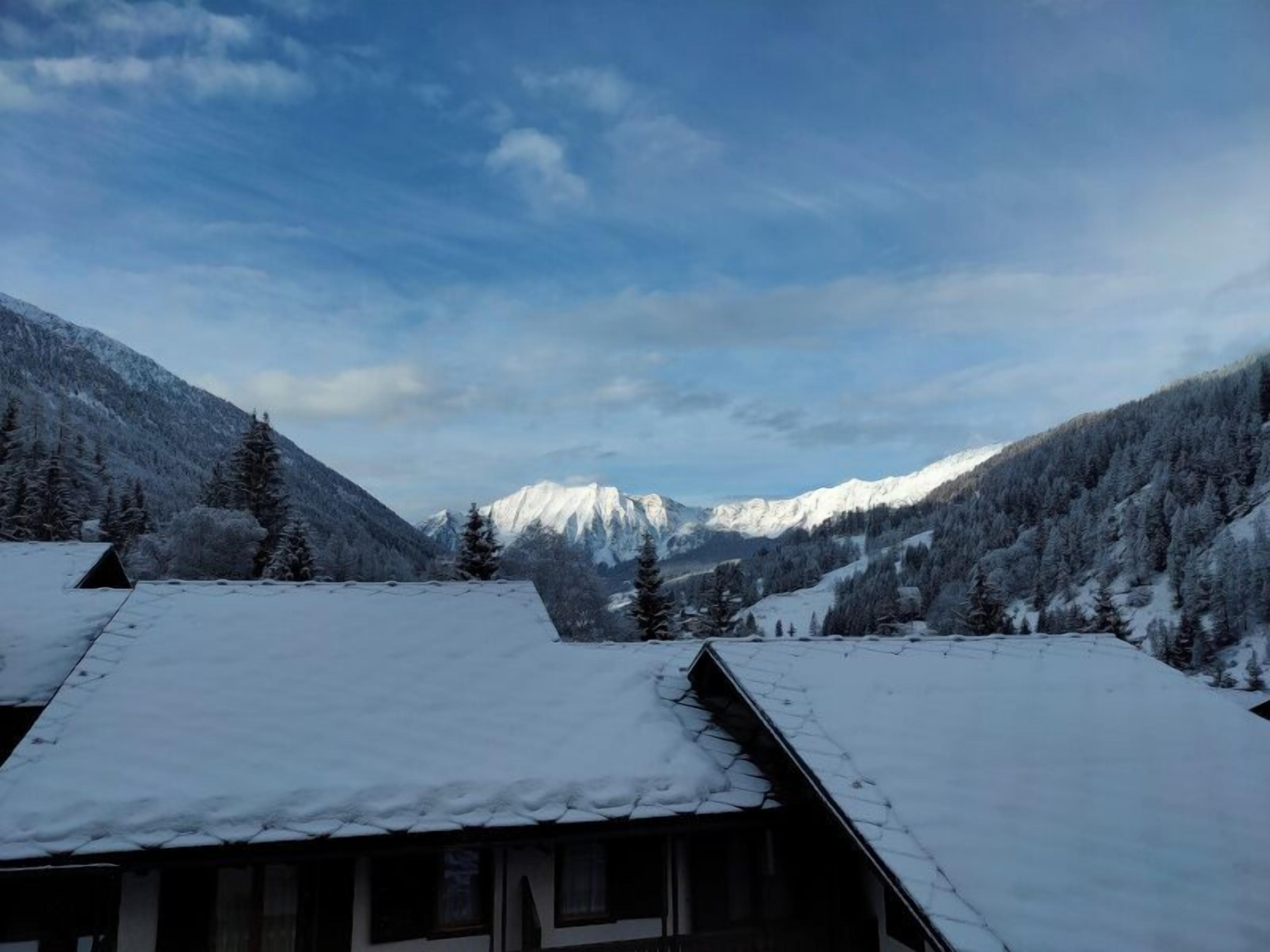 A cabin with a snow-covered roof, surrounded by a forested mountain landscape.
