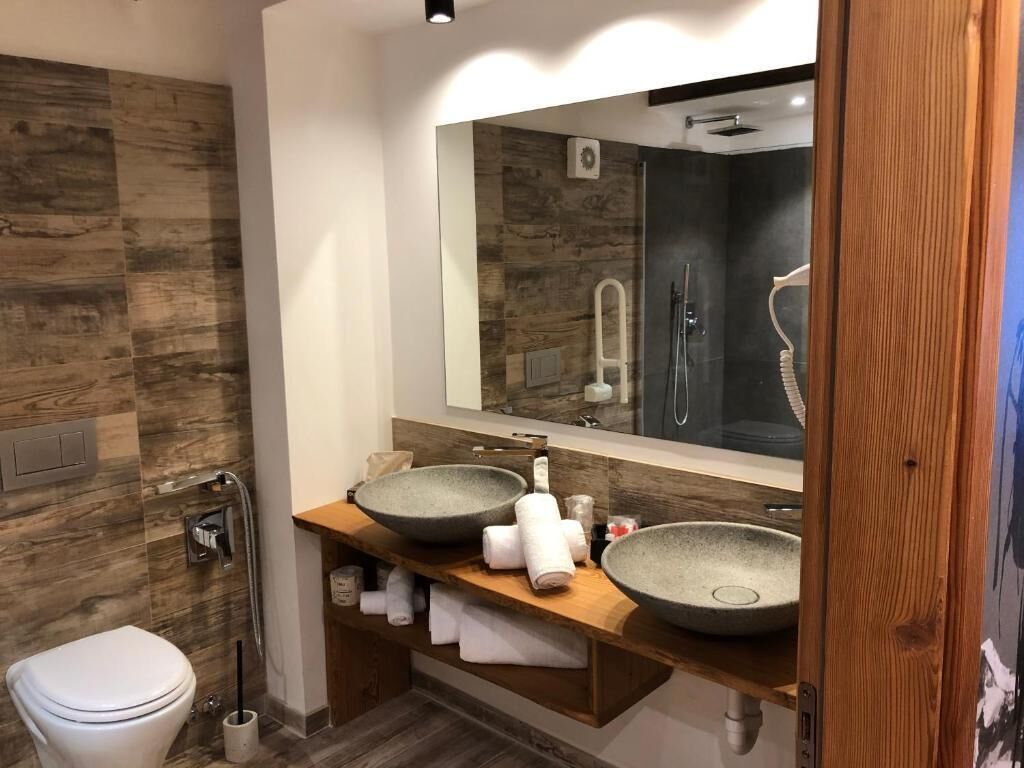 A modern bathroom with a wooden wall, a large mirror, two vessel sinks, a toilet, and a shower area.