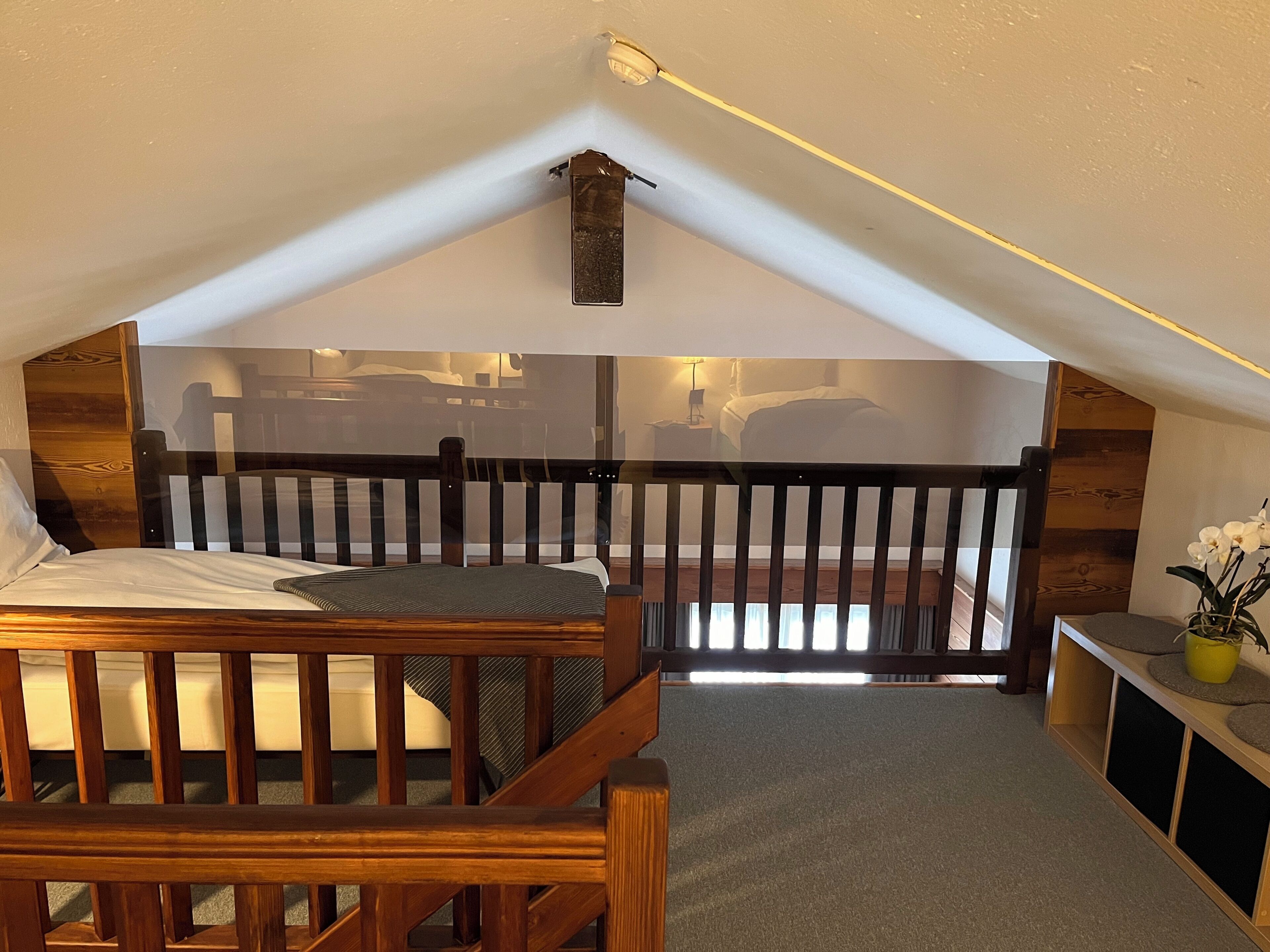 A small, cozy attic bedroom with a wooden bed, a small table, and a view of the living area below.