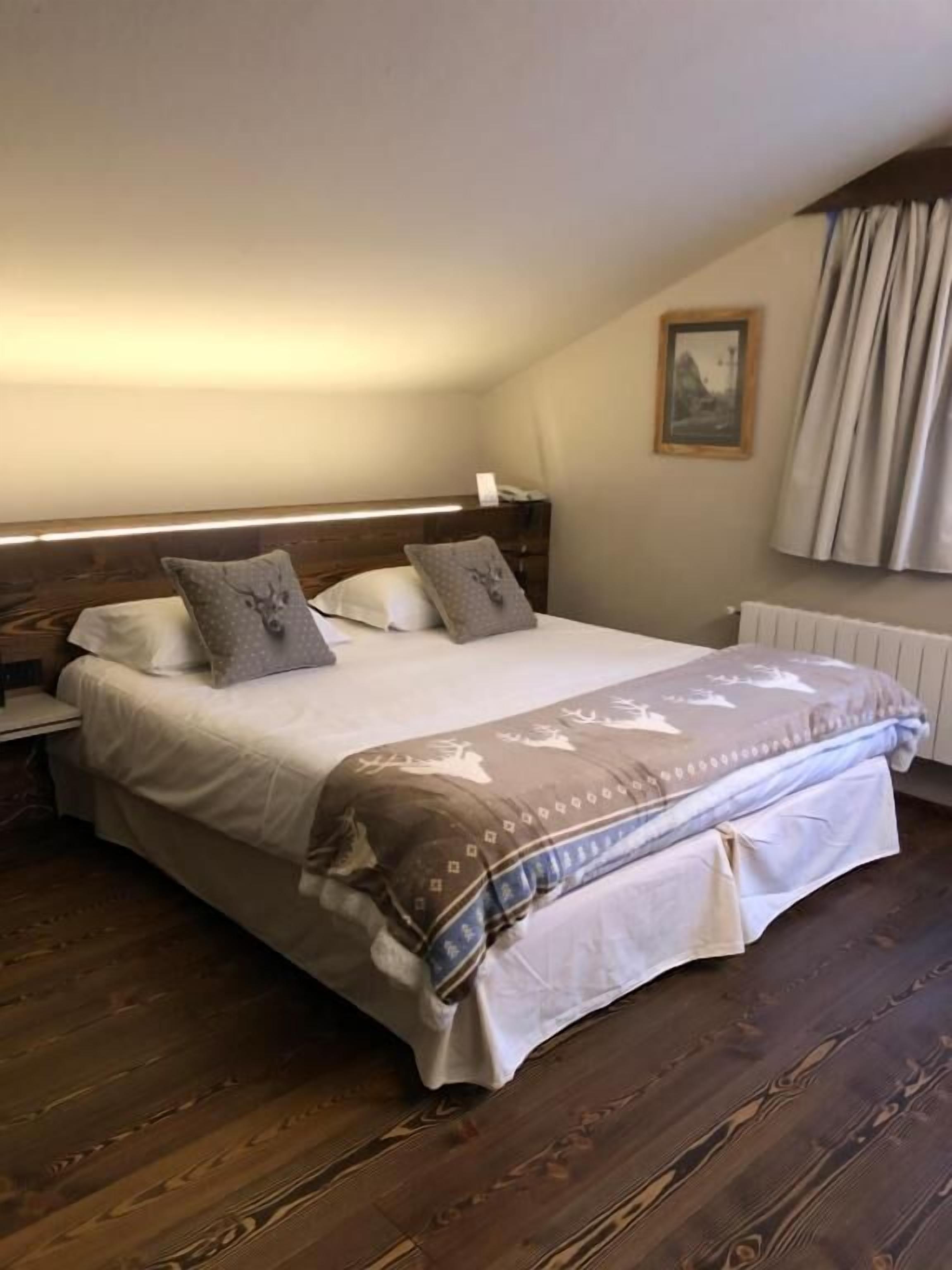 A bedroom with a wooden floor, a bed with a patterned bedspread, a nightstand, a wall-mounted picture, and a window with curtains.