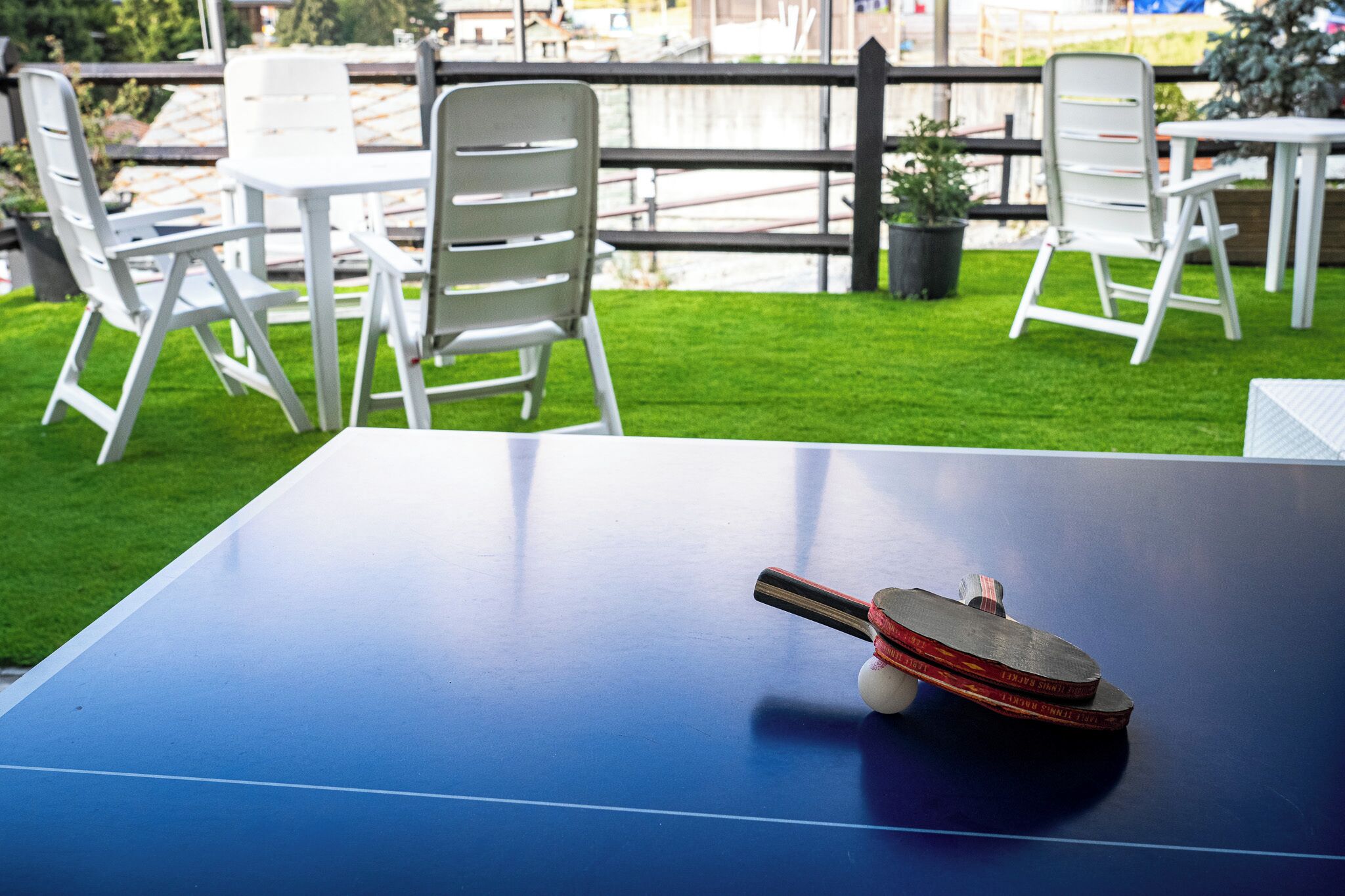 Una mesa de ping pong con una paleta y una pelota, rodeada de sillas blancas y un pequeño área de césped.