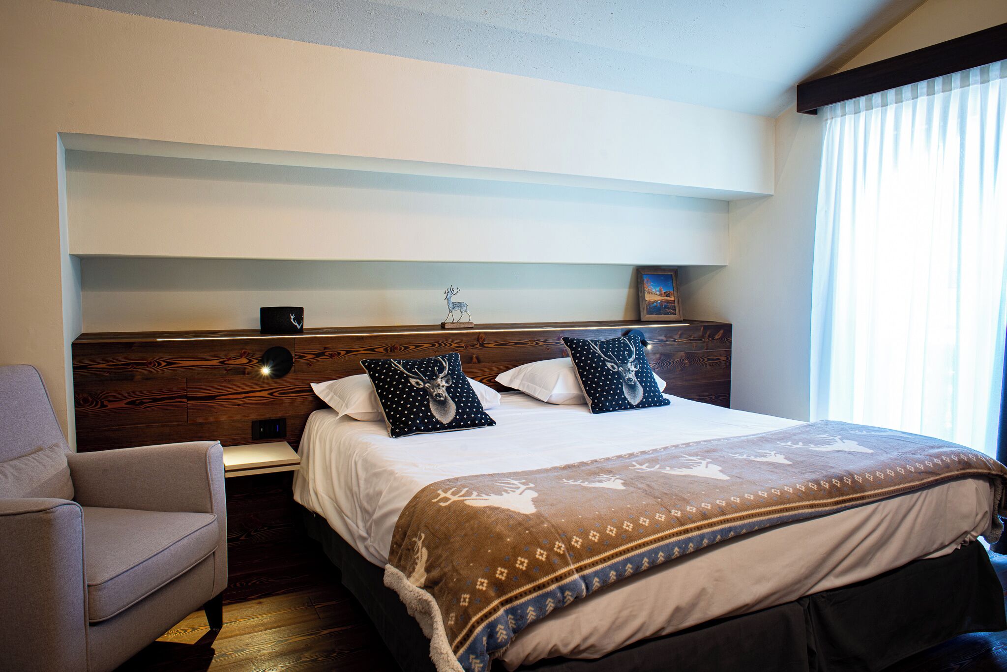 A hotel room with a wooden headboard, a bed with patterned bedding, a nightstand, a chair, and a window with sheer curtains.