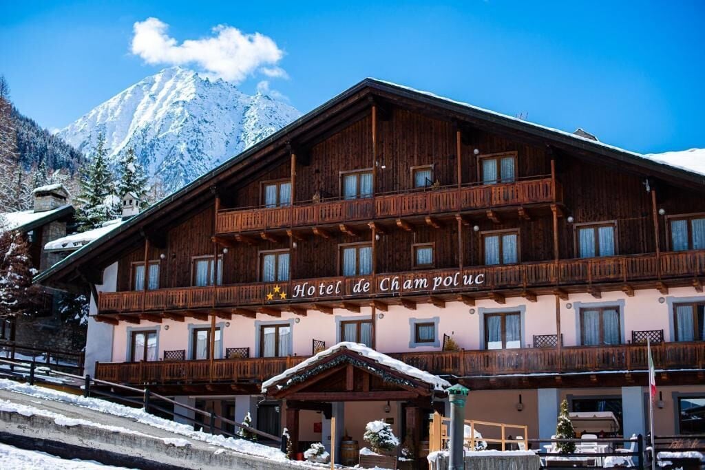Ein traditionelles Holzgebäude mit Balkonen, beschriftet 'Hotel de Cham poluC', vor einer verschneiten Bergkulisse.
