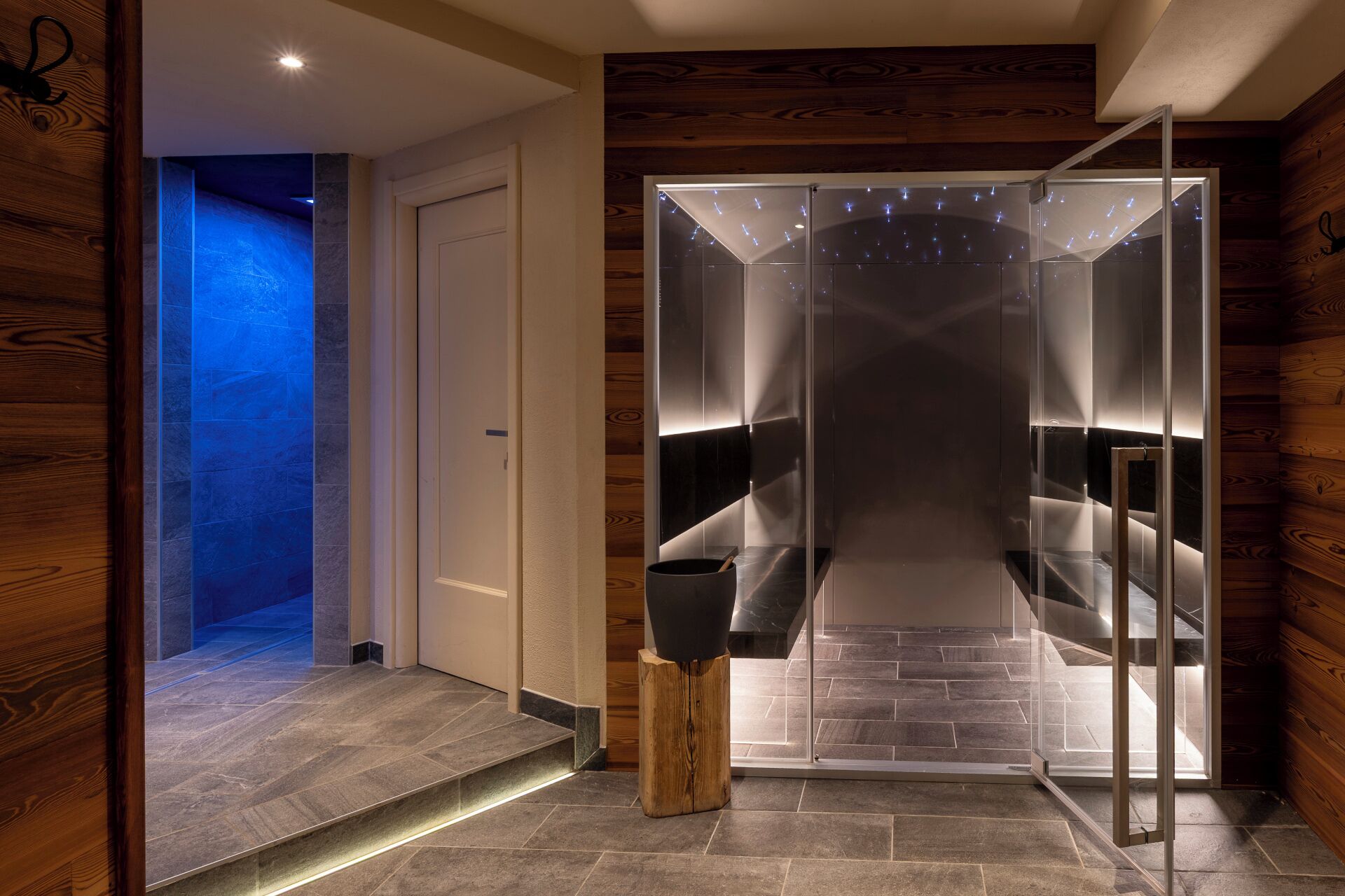 Un intérieur moderne avec un ascenseur en verre, un plancher en bois et une porte donnant sur un espace éclairé de lumière bleue.