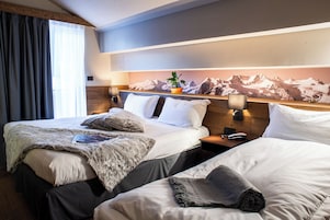 Habitación de hotel con dos camas, un mural con vistas a la montaña, una planta en la cabecera y una mesita de noche con lámpara.