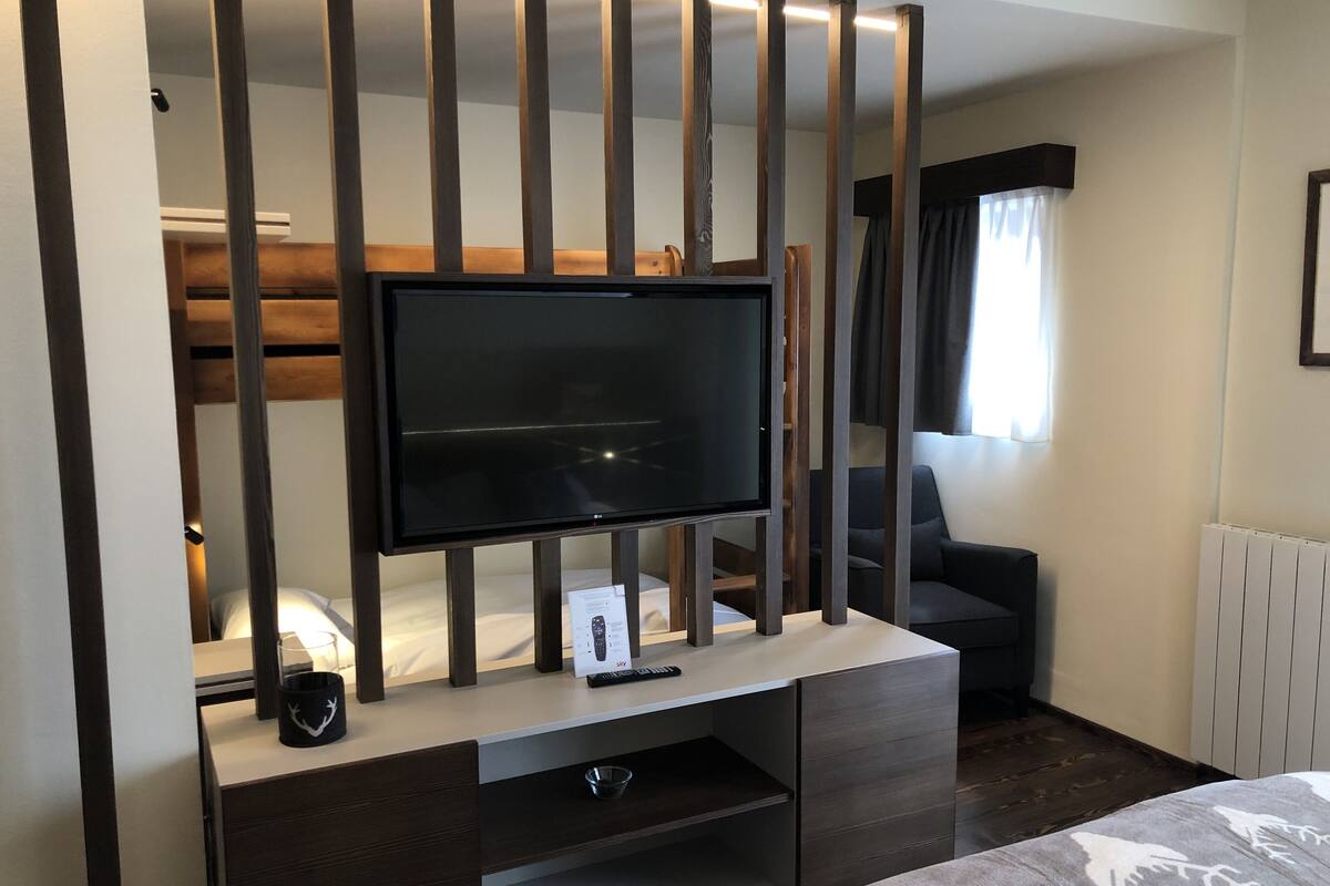 A modern hotel room with a flat-screen TV on a wooden stand, a sofa, a bed with a patterned bedspread, and a window with curtains.