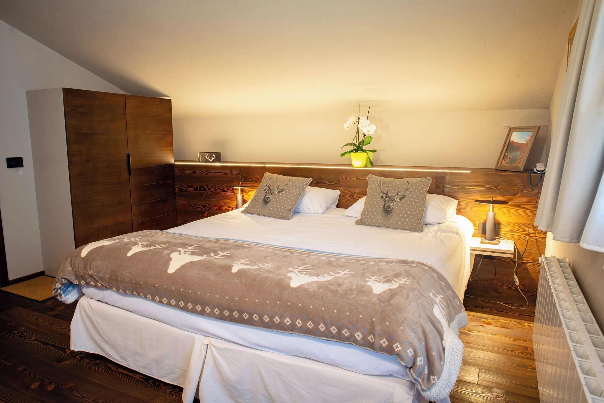 Une chambre à coucher avec un grand lit, une tête de lit en bois, des tables de chevet, une petite table, une plante en pot et un tableau encadré au mur.
