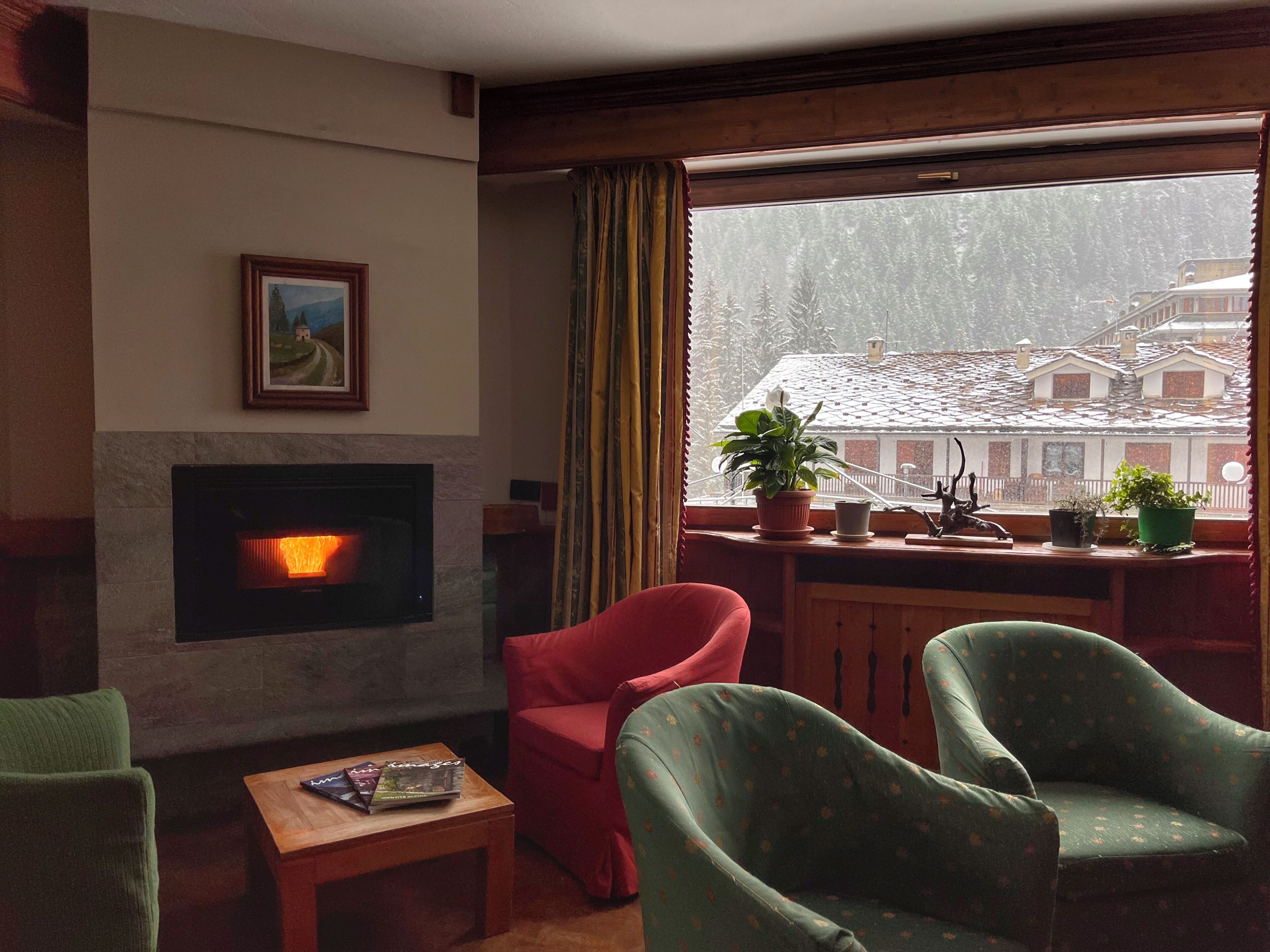 Una sala de estar acogedora con chimenea, un sillón rojo, sillones verdes y vistas a un paisaje nevado a través de una ventana.