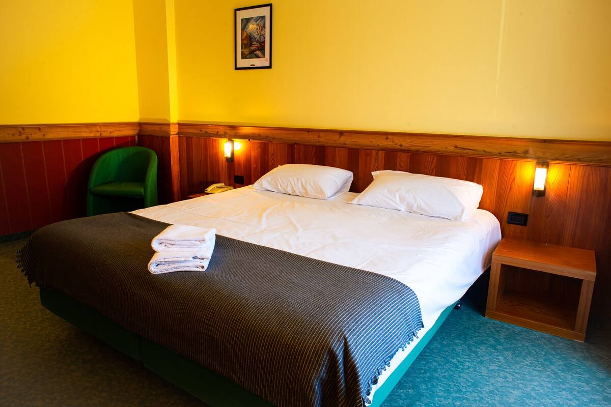 A hotel room with a bed, a green chair, a wooden headboard, and two wall-mounted lights.