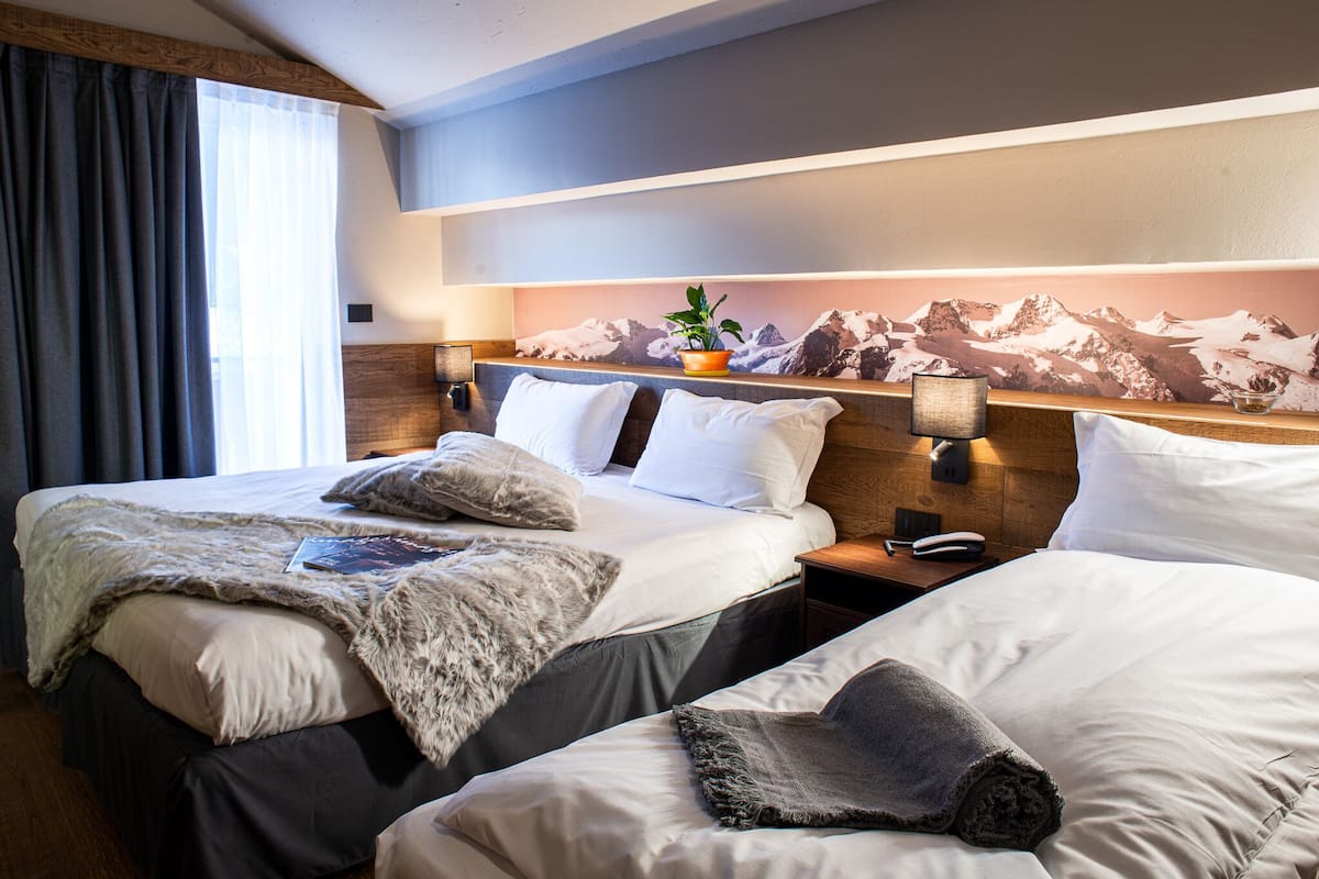 A hotel room with two beds, a mountain view mural, a plant on the headboard, and a bedside table with a lamp.