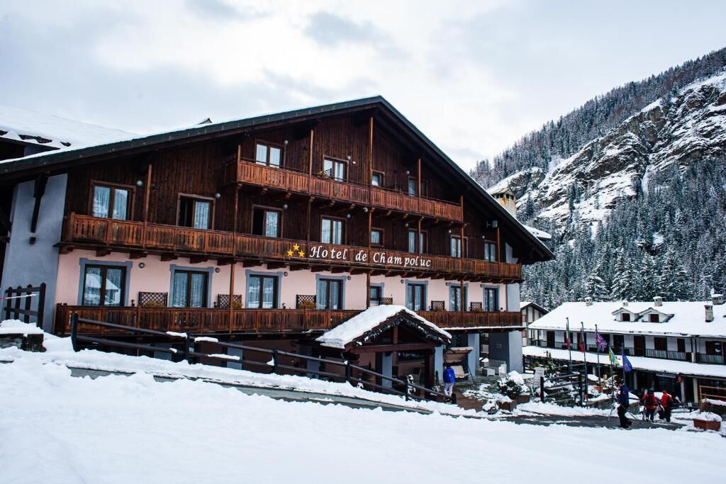 Et hotel med flere etager og balkoner, en skibakke i forgrunden og snedækkede bjerge i baggrunden.