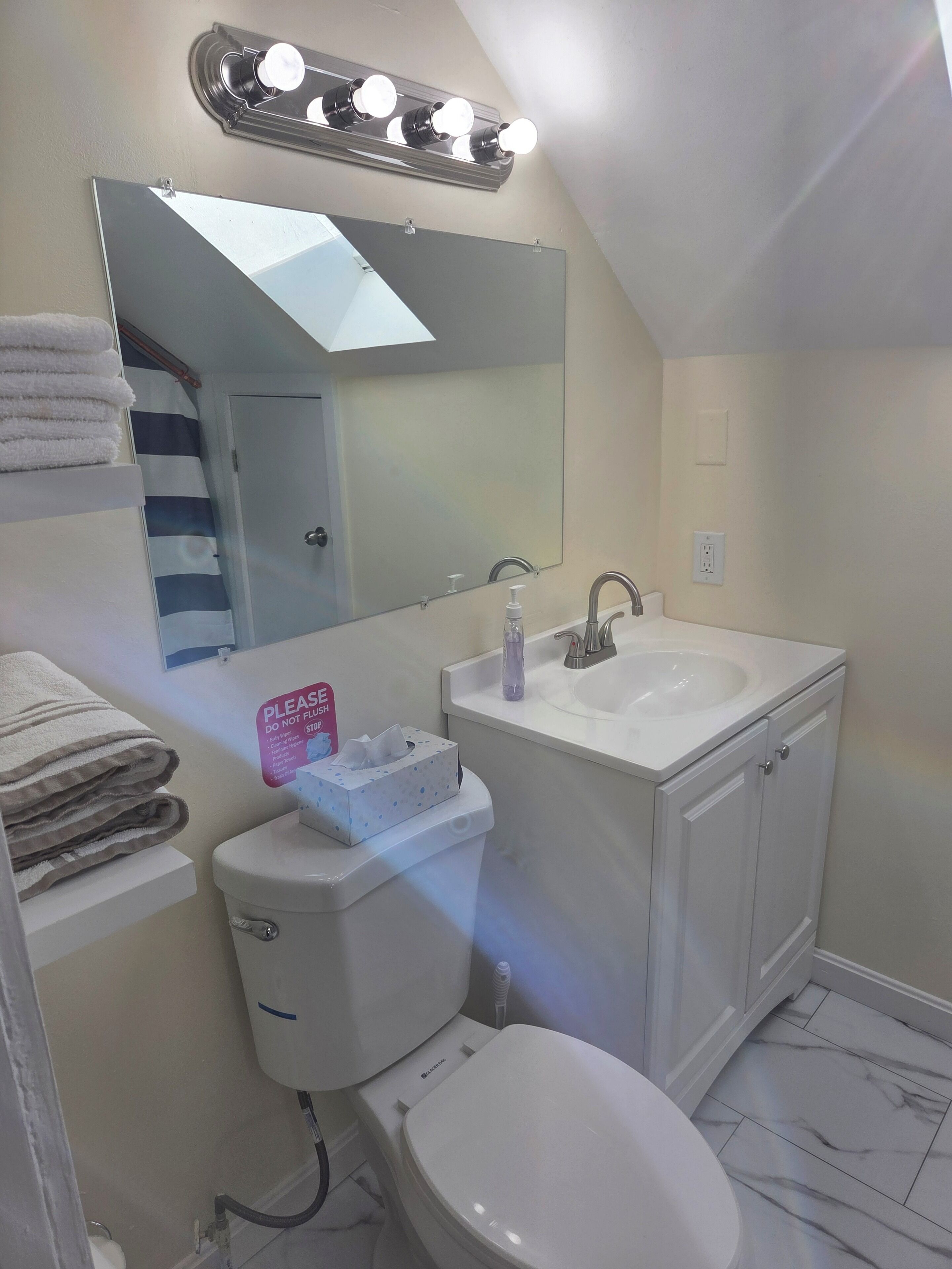 Un baño con inodoro, lavabo y espejo. Toallas apiladas en un estante, y un cartel en la pared.