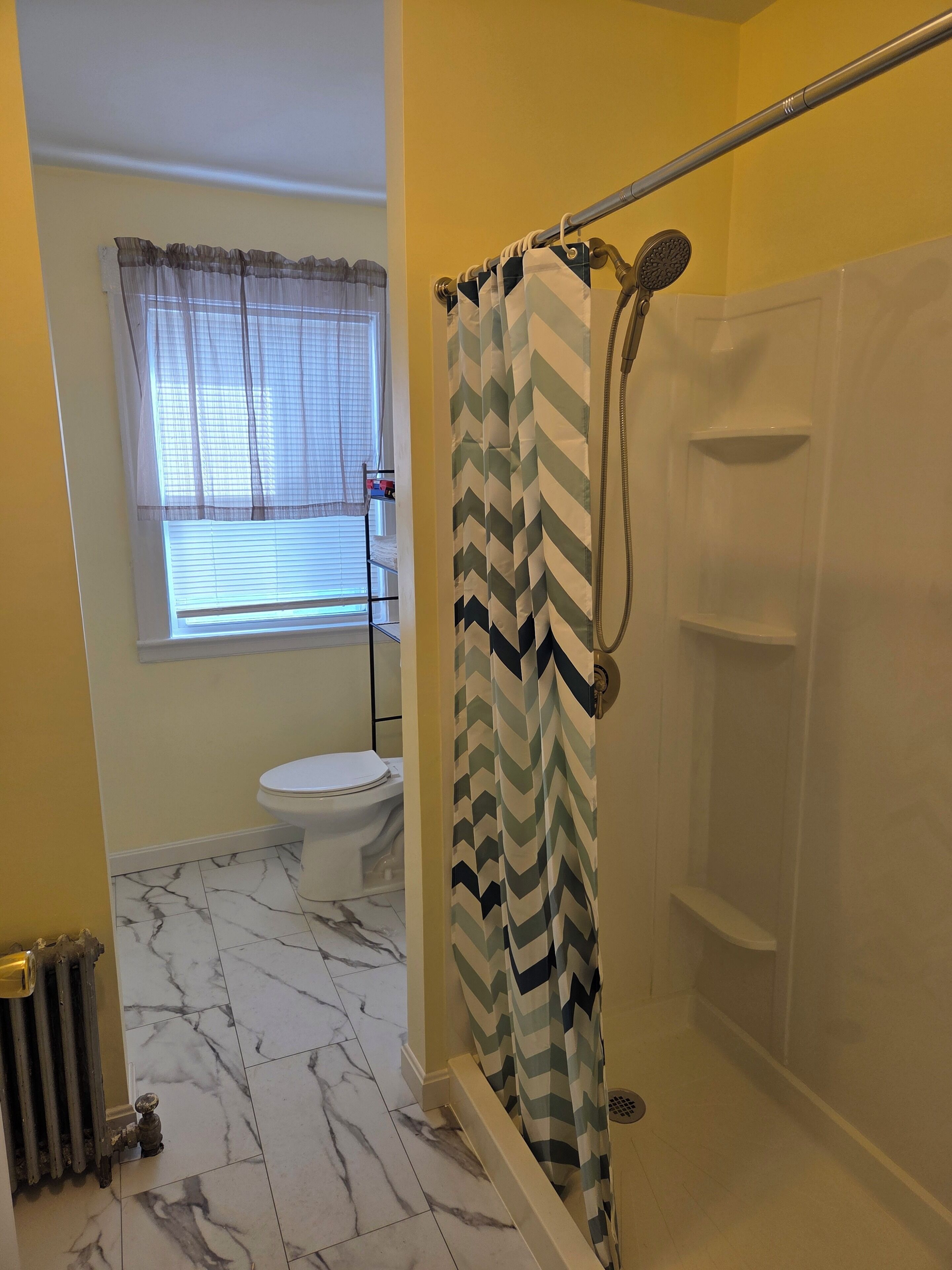 A bathroom with a shower, toilet, and a window with blinds.