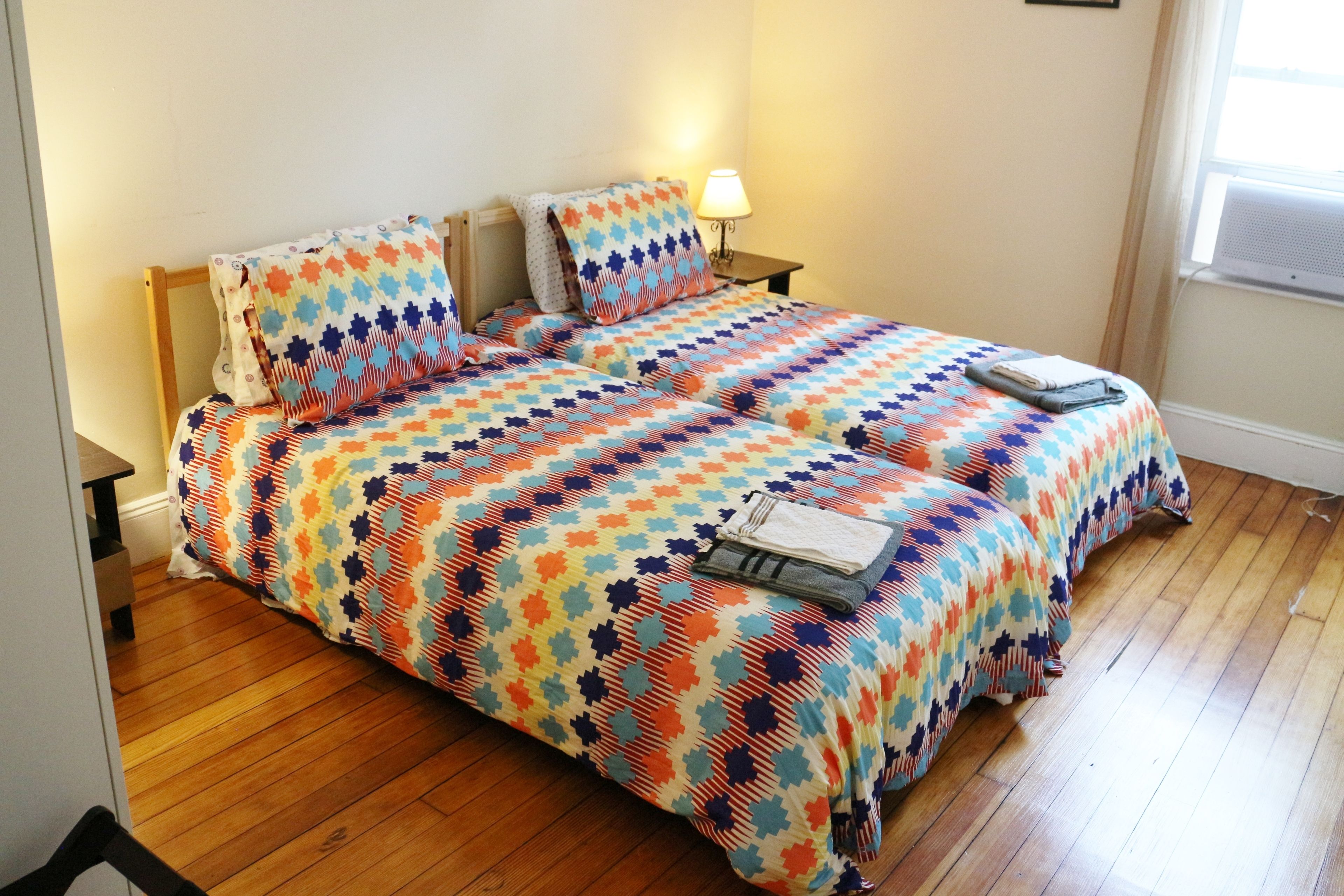A double bed with a colorful geometric-patterned bedspread, a bedside table with a lamp, and a window with an air conditioning unit.