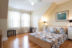 A bedroom with a bed, a desk, a chair, a ceiling fan, and a painting on the wall.