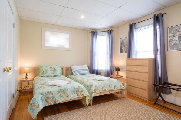 A bedroom with two single beds, a wooden dresser, a window with curtains, a framed picture on the wall, and a small table with a lamp.