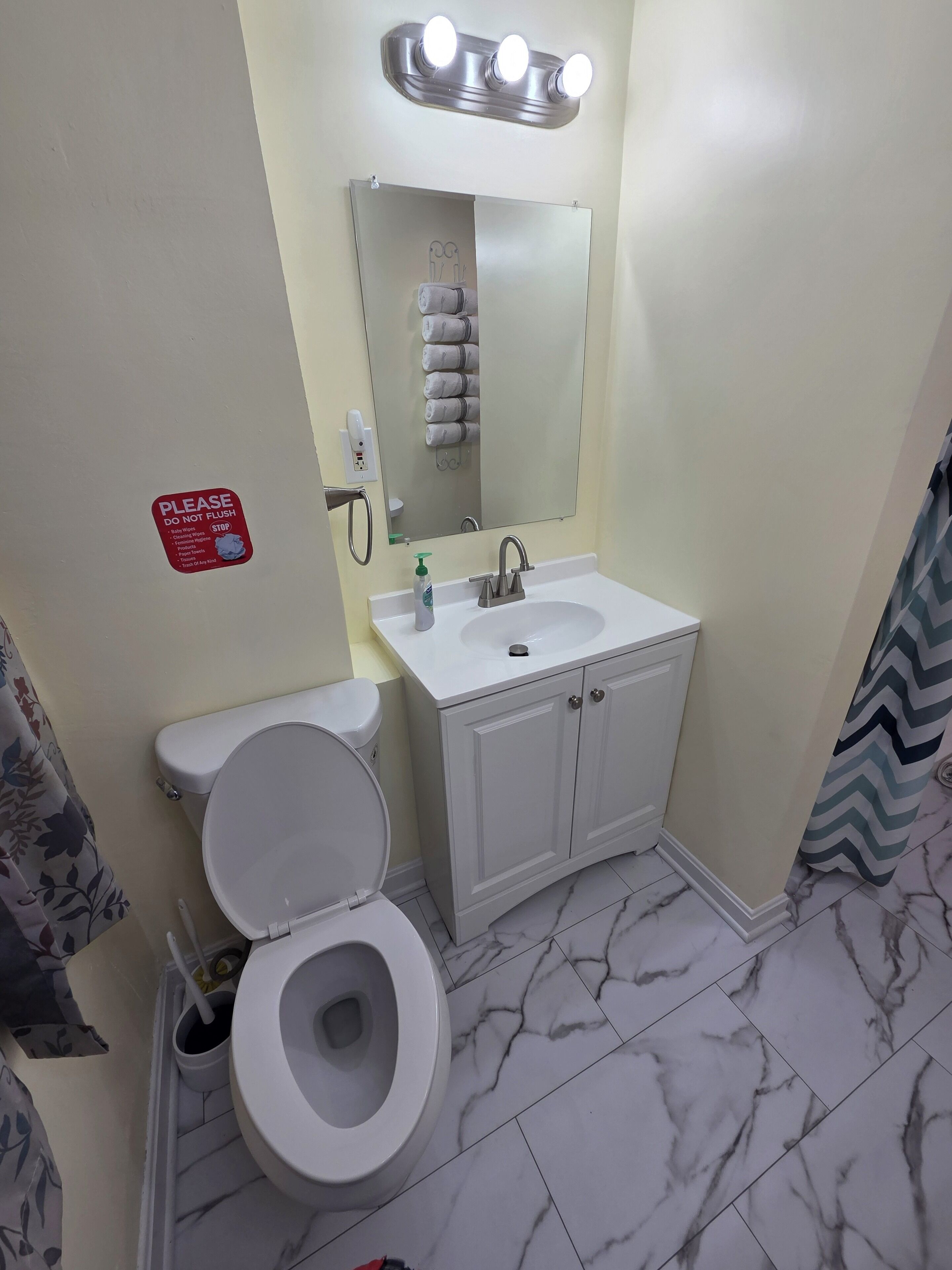 Un baño con inodoro, lavamanos y espejo. Hay un letrero en la pared y una cortina de ducha.