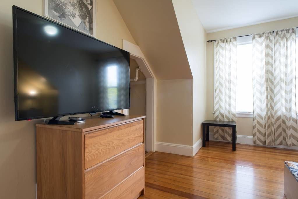 Una habitación con una televisión de pantalla plana sobre un tocador de madera, una mesita y una ventana con cortinas a cuadros.