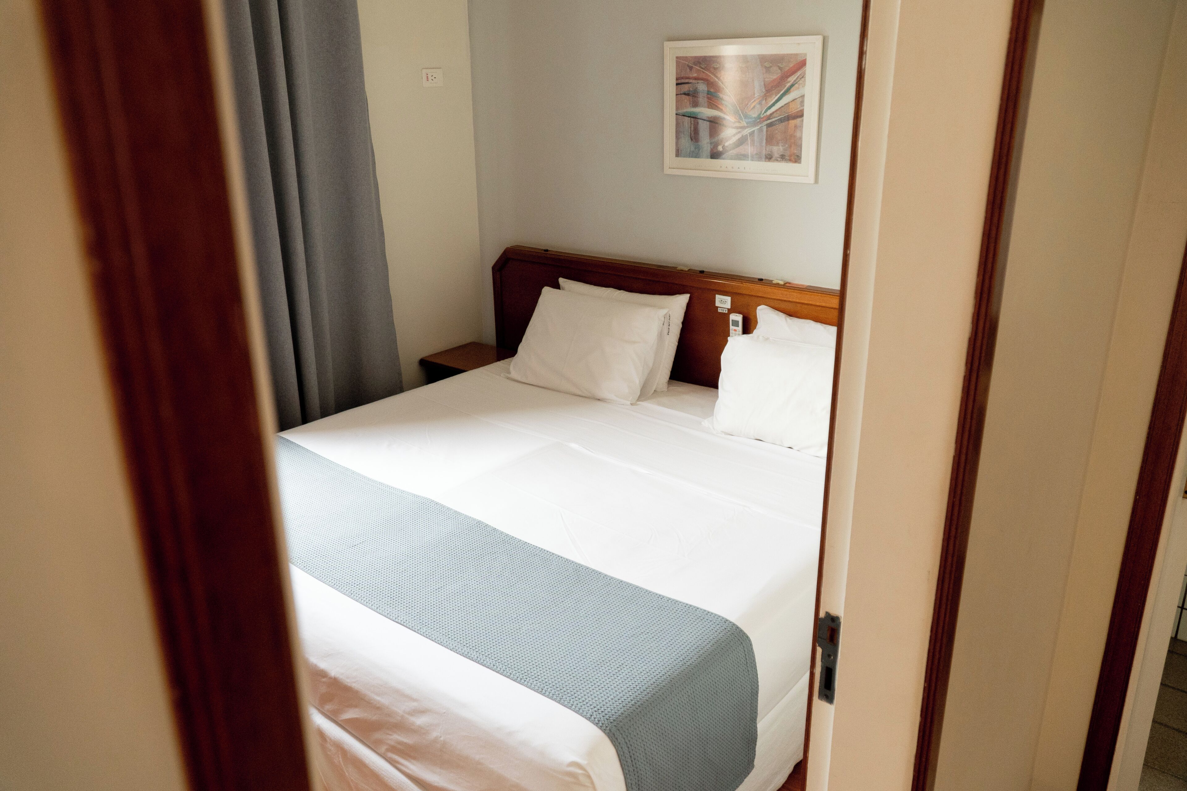 Una habitación de hotel con cama, mesita de noche, un cuadro en la pared y ventana con cortinas.