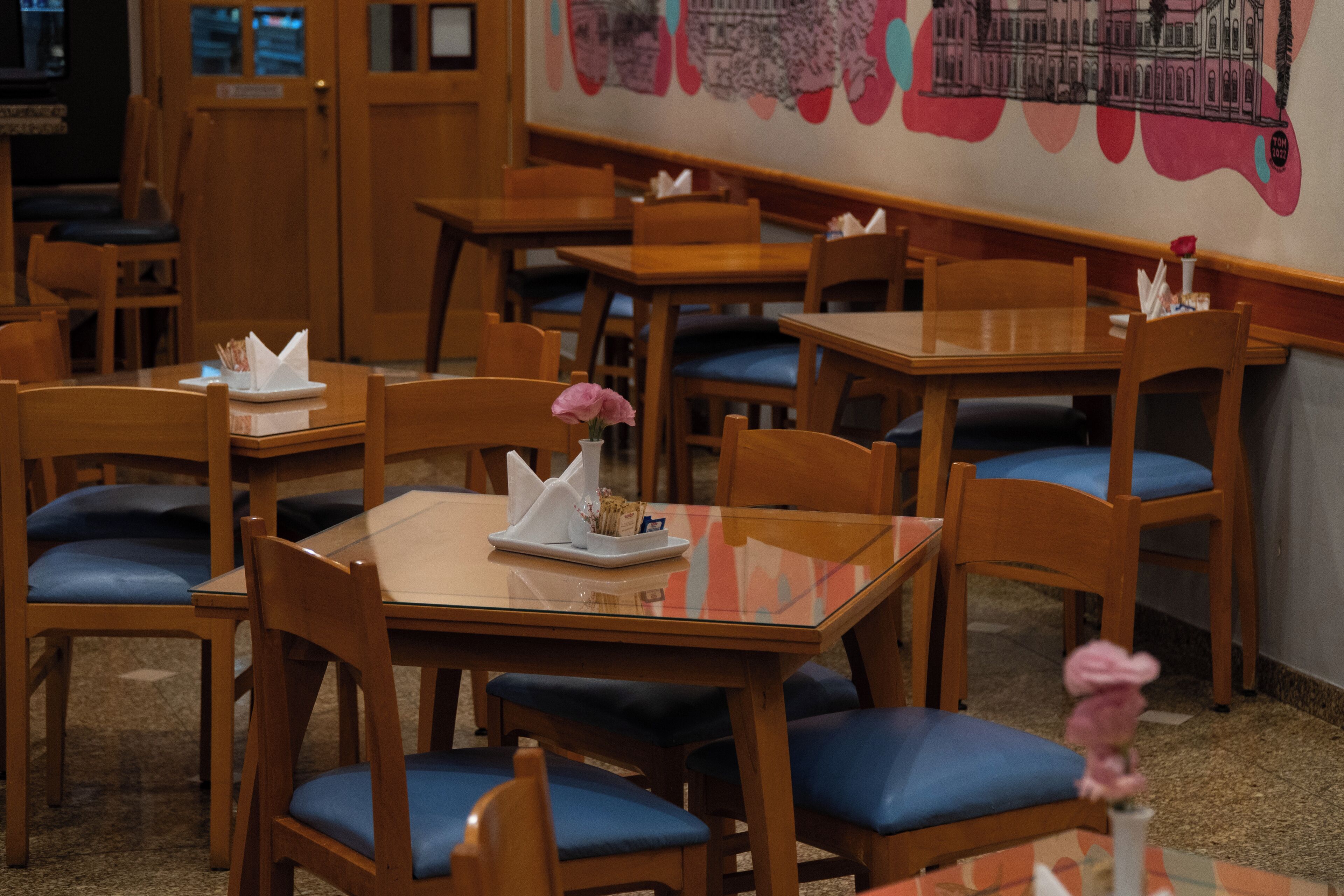 A restaurant interior with wooden tables and blue cushioned chairs, some with napkins and a vase of flowers.