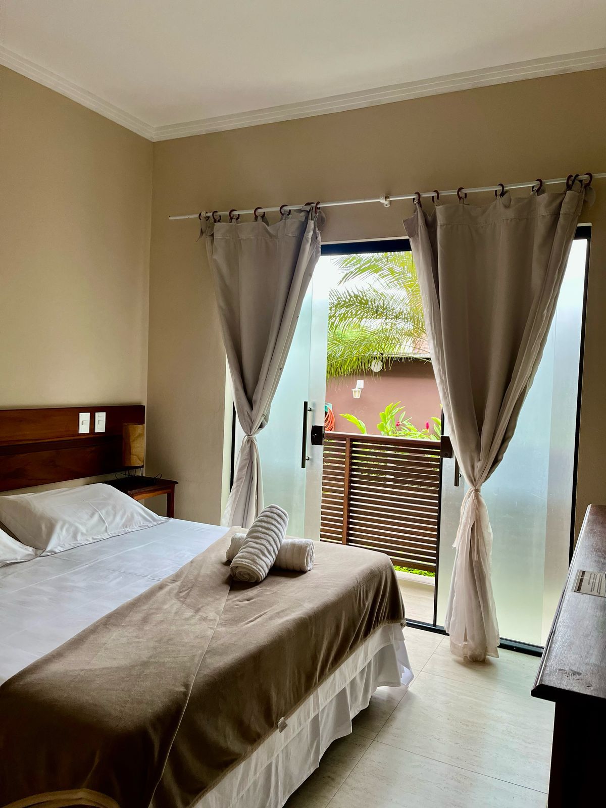 A bedroom with a bed, curtains, a nightstand, and a view of greenery outside.