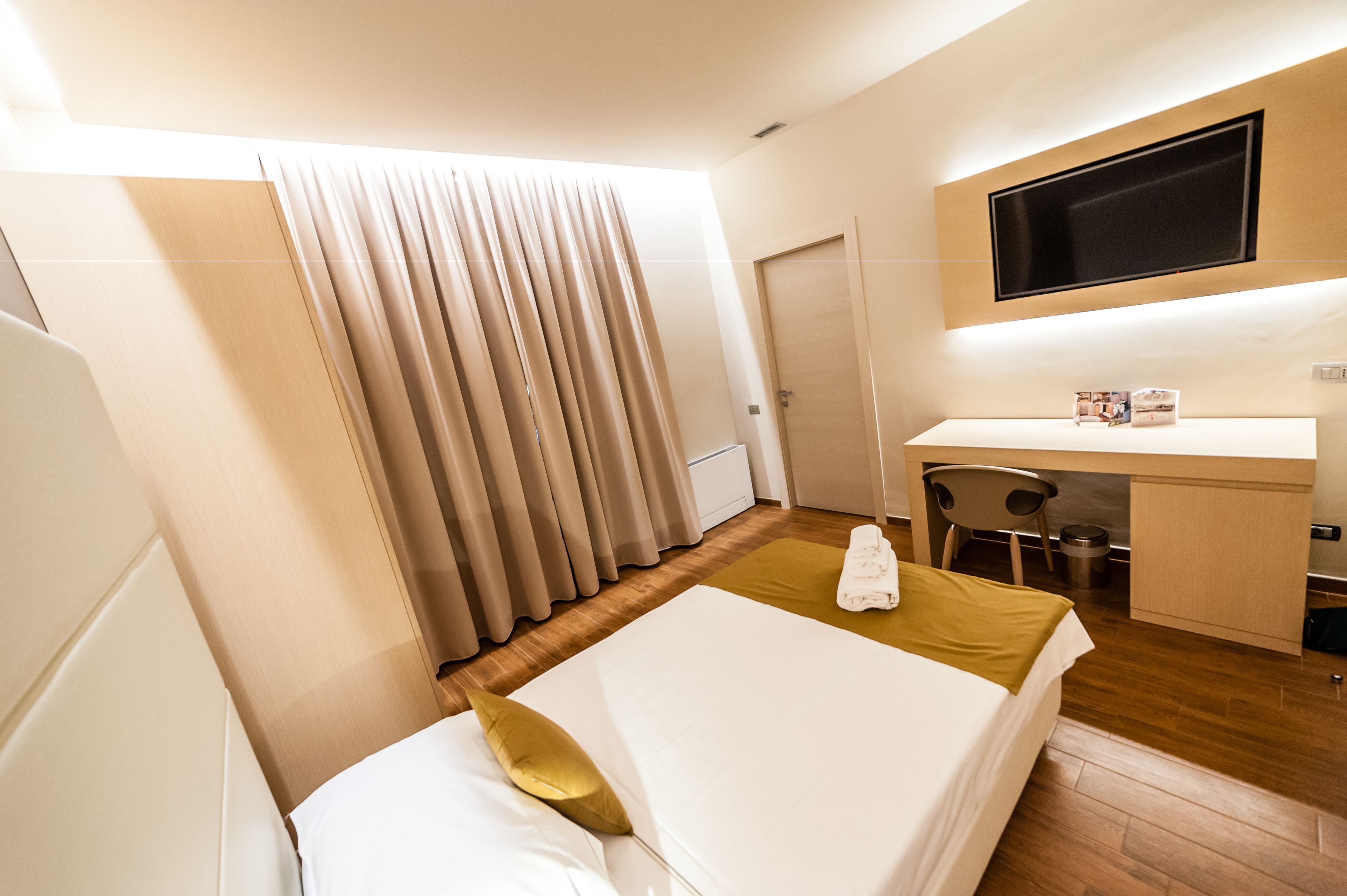 A hotel room with a bed, a desk, a chair, a TV mounted on the wall, and a window with curtains.