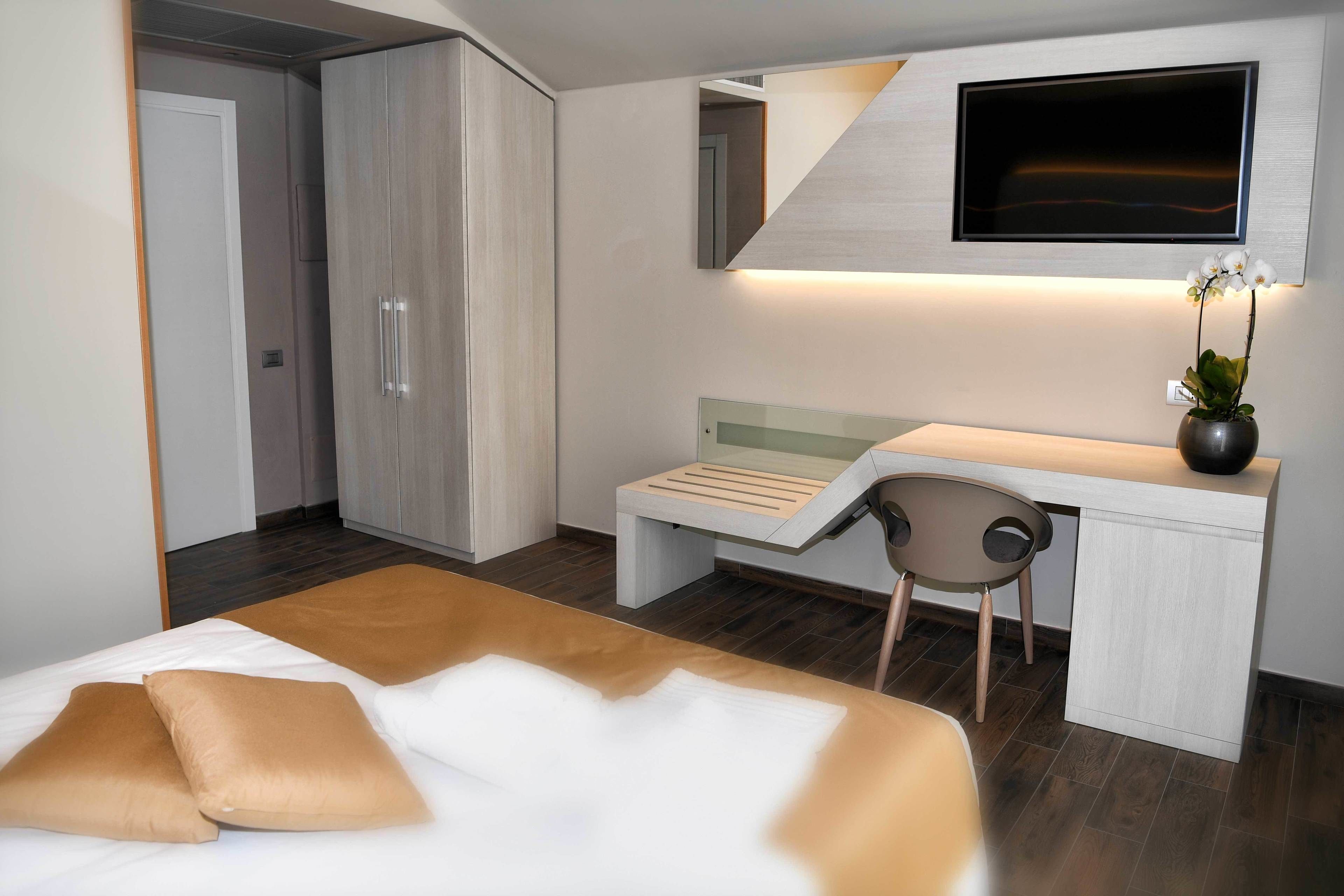 A modern hotel room with a bed, a desk, a chair, a TV, and a wardrobe.