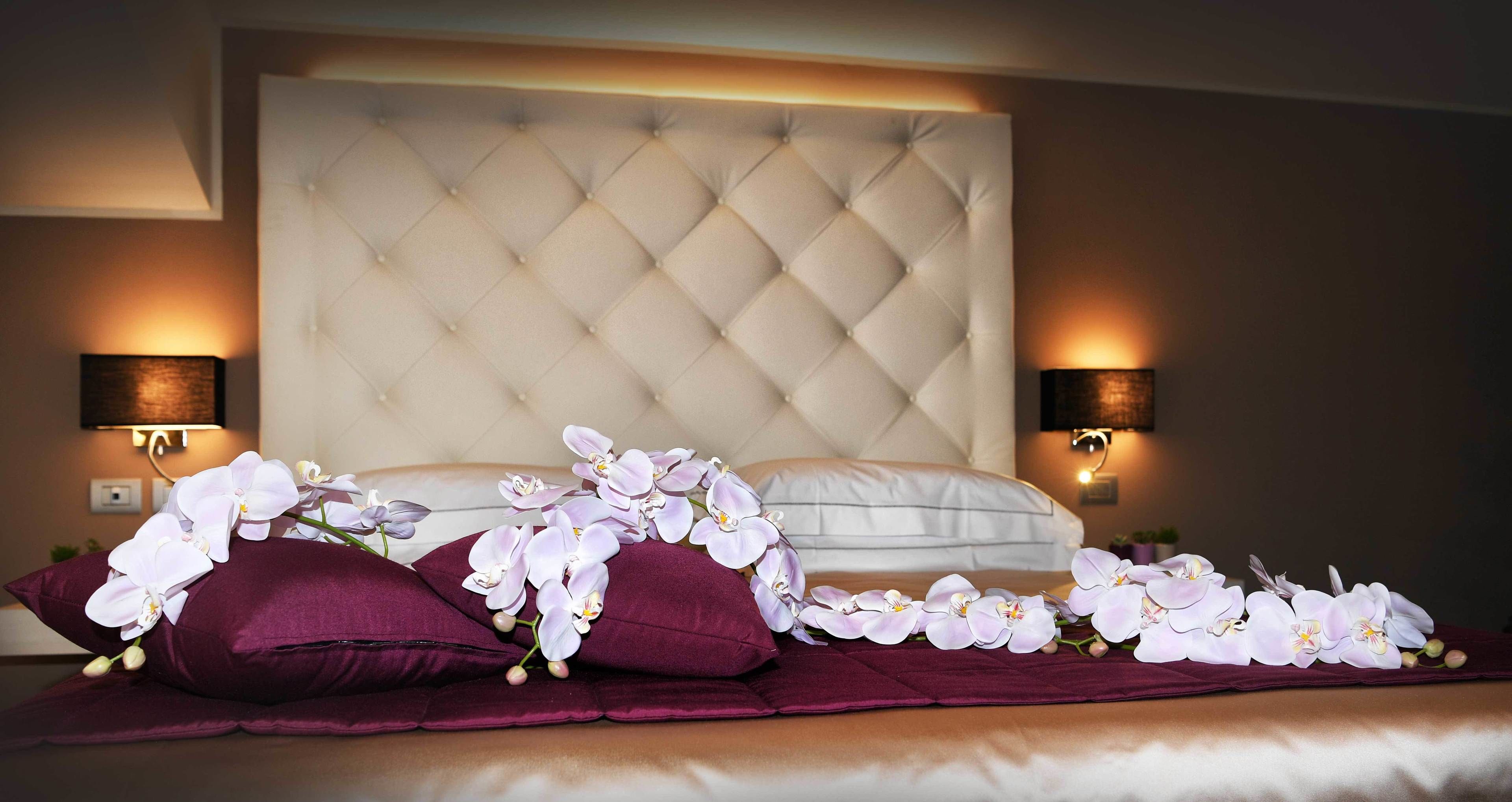 Una cama bien tendida con un cabecero capitonado, dos lámparas fijadas a la pared y un arreglo decorativo de orquídeas.