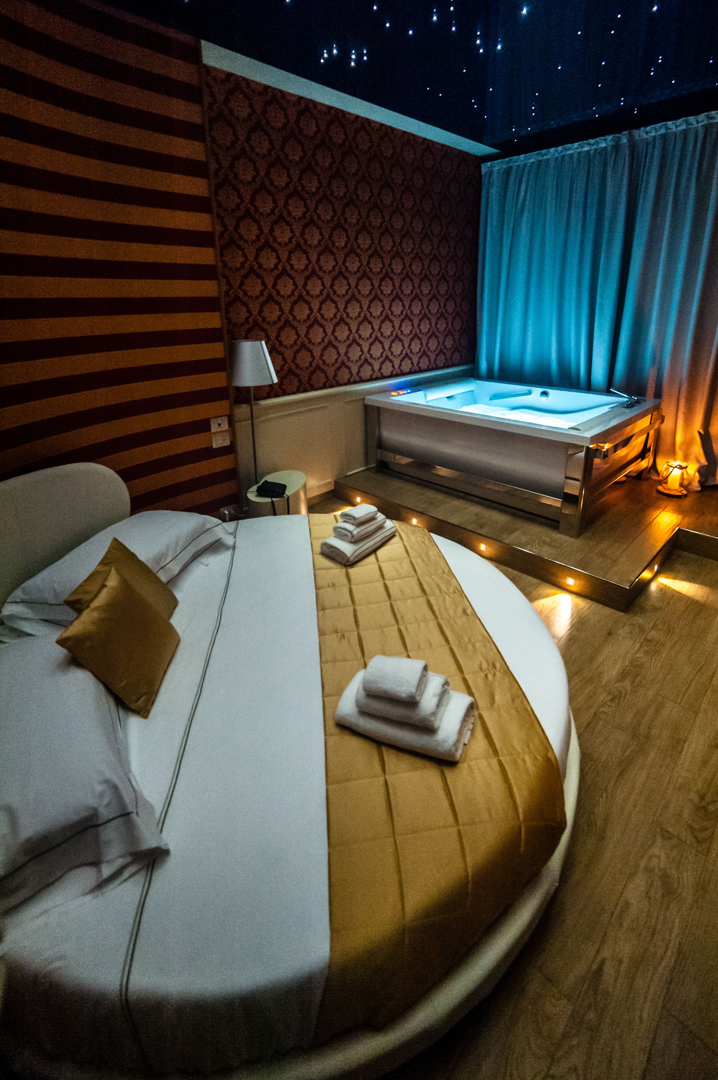 A hotel room with a bed, a small table, a nightstand, a window with curtains, and a wall with a patterned design.