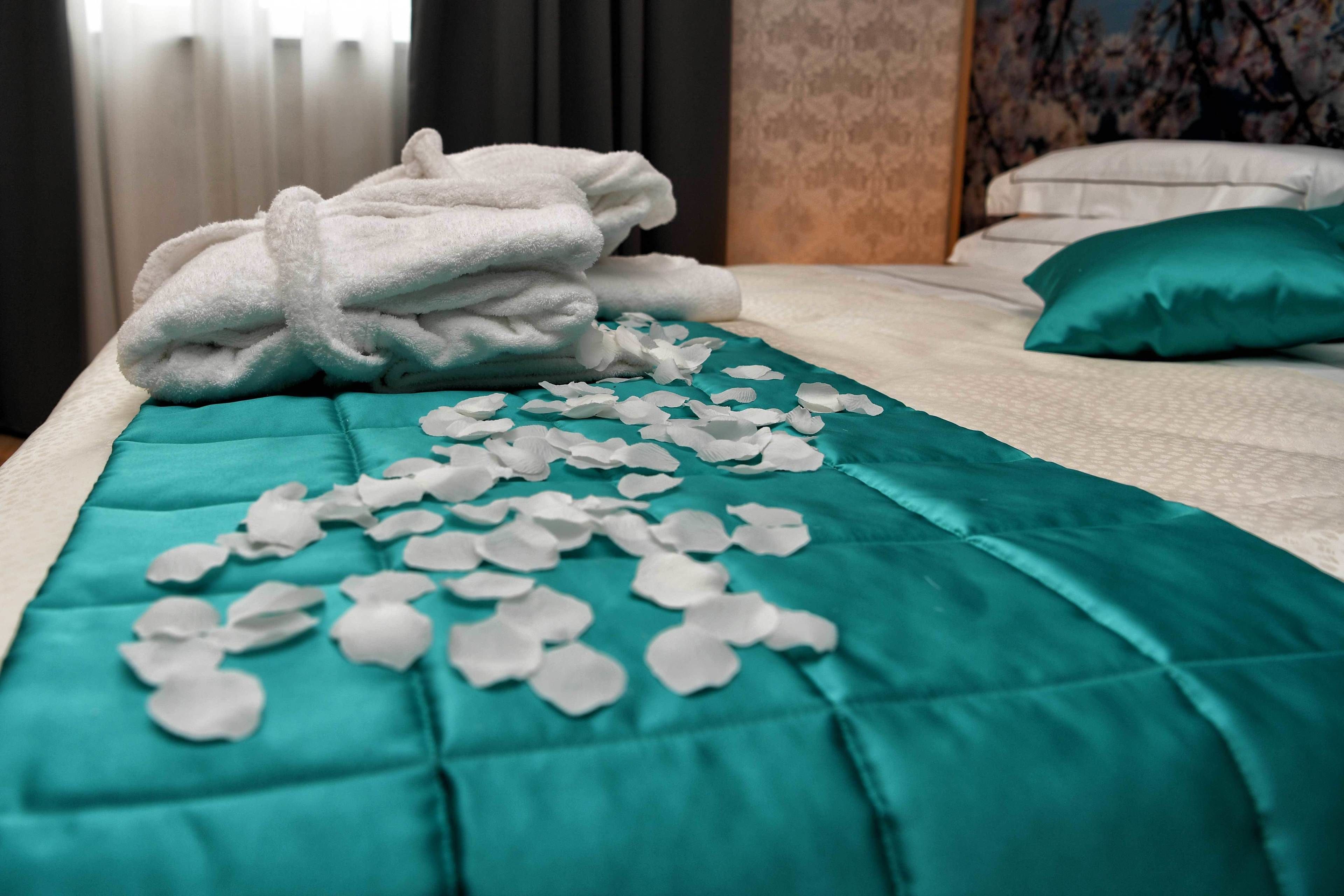 A neatly made bed with a teal bedspread, white pillows, and a stack of white towels.