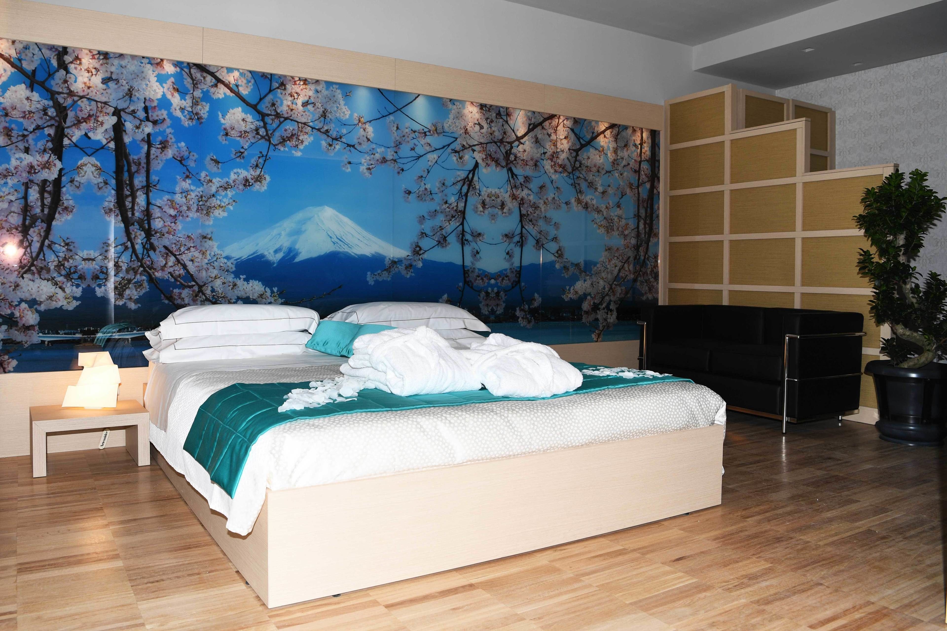 A bedroom with a bed, a nightstand, a large wall mural, a black sofa, and a potted plant.