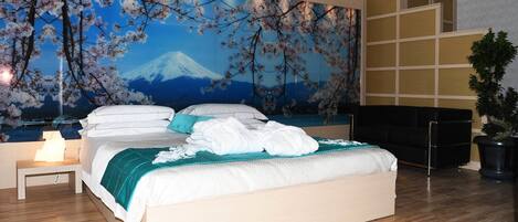 A bedroom with a bed, a nightstand, a large wall mural, a black sofa, and a potted plant.