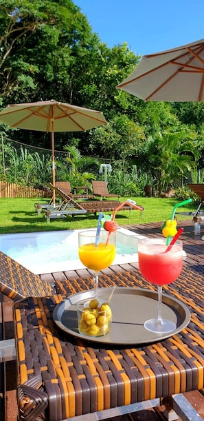 Escena junto a la piscina con dos bebidas sobre una mesa, rodeada de sillones de descanso y sombrillas.