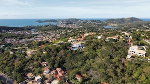 Un pueblo costero con edificios residenciales y vegetación, una casa azul destacada con piscina, y vistas al mar y colinas al fondo.