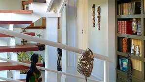 A modern interior with a staircase, a round table with a red vase, and a bookshelf with various decorative items. - Top de Buzios Boutique (Búzios)