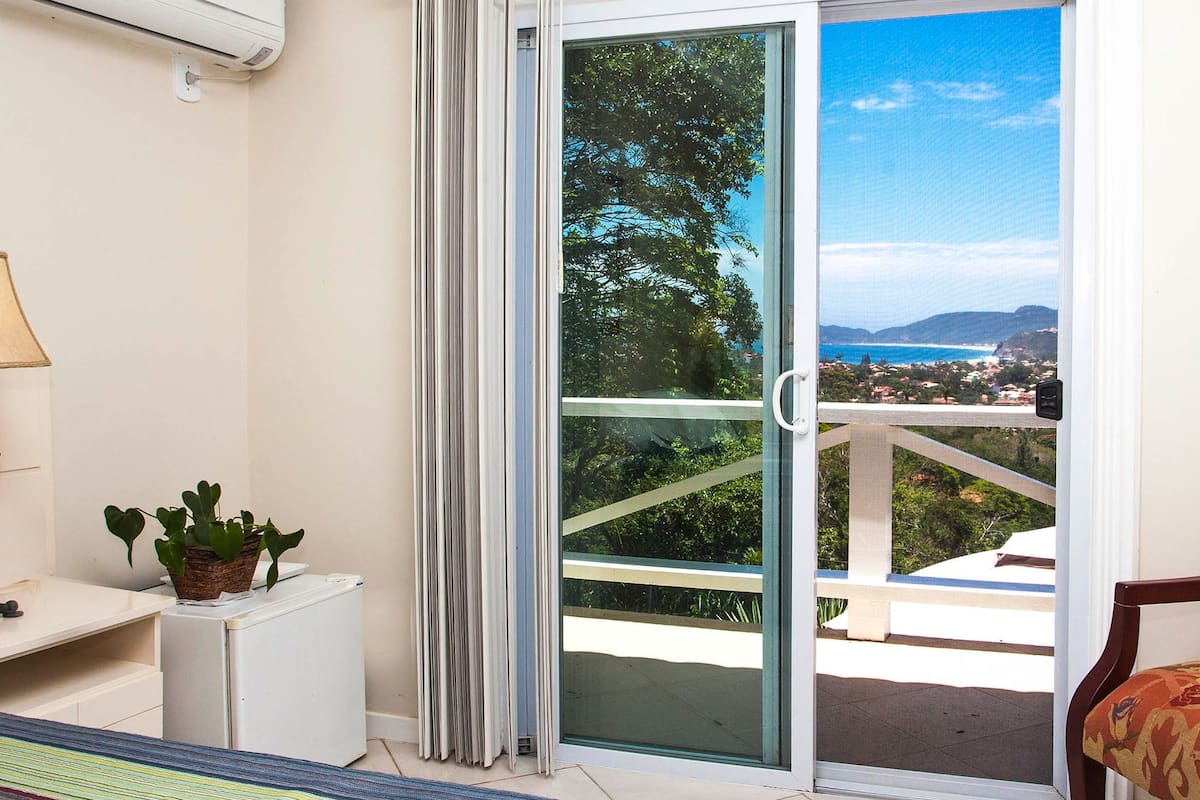 Une chambre d’hôtel avec un lit, une petite table avec une lampe, une plante en pot et une porte-fenêtre coulissante donnant sur un balcon avec vue sur la mer et les collines.
