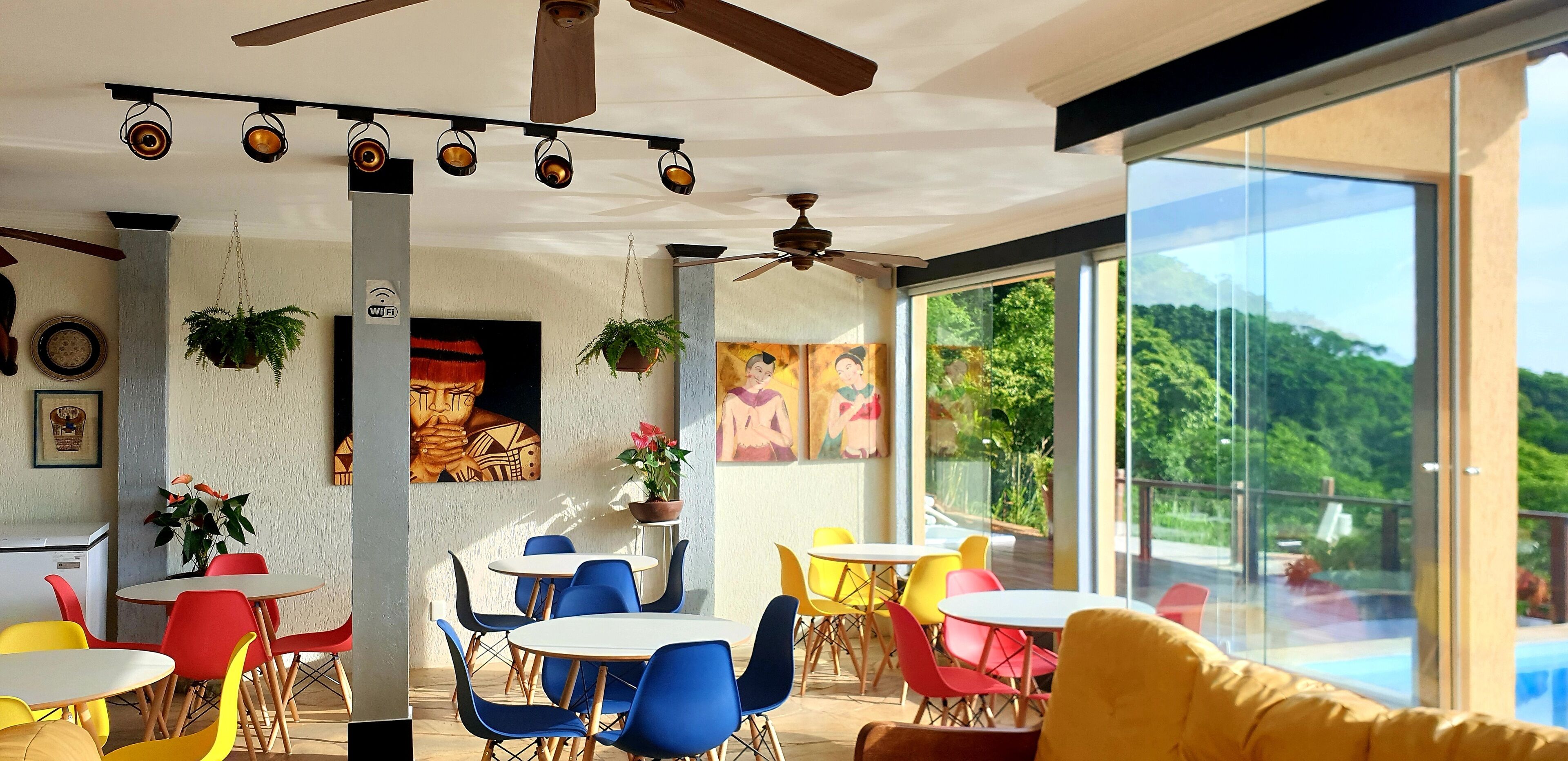 Uma área de jantar moderna com mesas redondas e cadeiras coloridas, amplas janelas com vista para o verde e ventiladores de teto.