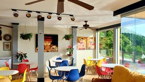 A modern dining area with round tables and colorful chairs, large windows with a view of greenery, and ceiling fans. - Top de Buzios Boutique (Búzios)