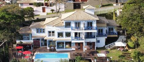 Una amplia casa de dos pisos con piscina, rodeada de vegetación exuberante y colinas al fondo.
