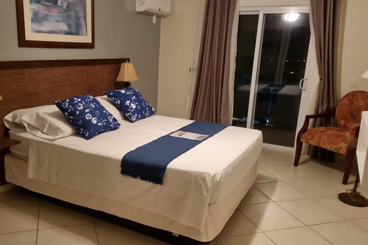 Un lit bien fait, avec une couvre-lit bleue et des oreillers, une table de chevet en bois avec une lampe et un téléphone, une chaise, et une vue sur l’extérieur à travers une porte vitrée.