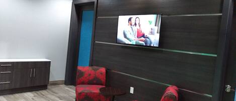 Una habitación con pared de madera oscura, un televisor de pantalla plana sobre un sofá y dos sillas rojas con estampado floral.